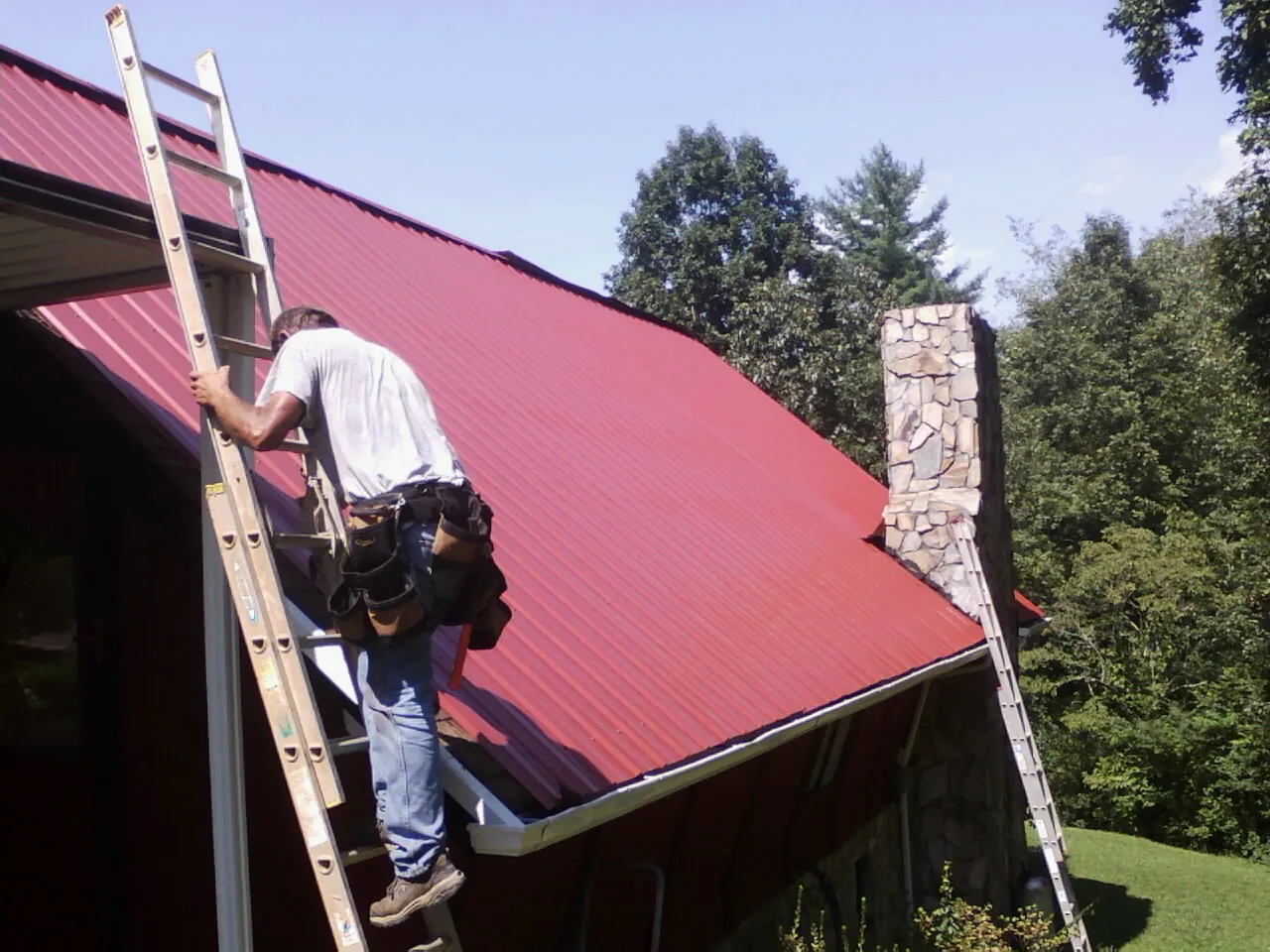 Roofer on a ladder beside a red metal roof and stone chimney — Soffit & Fascia Repair by Roofing Brothers in Heislerville